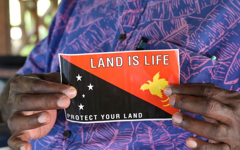 Eine indigene Frau hält die Flagge von Papua-Neuguinea in die Kamera. Zu sehen sind nur die Flagge und die Hände der Frau.