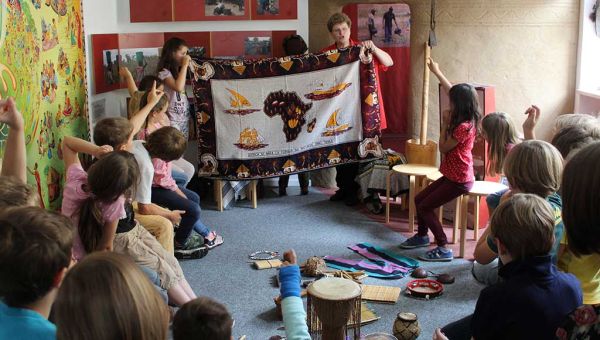 Eine Grundschulklasse zu Besuch bei missio München: Die Schülerinnen und Schüler sitzen im Ausstellungsraum in einem Kreis. Vorne zeigt eine missio-Mirarbeiterin ein großes Tuch mit einer gezeichneten Landkarte von Afrika.