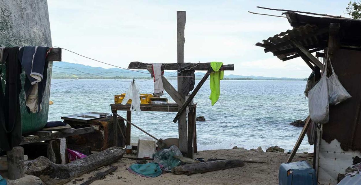 Blick von einer kleinen Insel in Ozeanien auf das Meer und den gegenüberliegenden Küstenabschnitt. Im Vordergrund sind im Sand ein einfaches Holzkreiz zu sehen, das mit Tüchern geschmückt ist. Rechts und links stehen einfache Hütten.