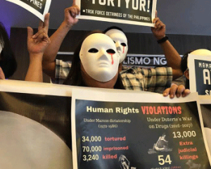 Protestierende Menschen mit Masken auf den Philippinen. Aus dem linken Auge der weißen Masken läuft Blut. Sie halten Plakate, auf denen die Menshenrechtsverletzungen und die hohe Zahl der Toten und Gefangenen angeprangert wird.