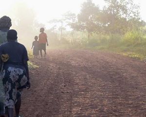 Auf der Insel Buvuma in Uganda laufen zwei Frauen auf einem am Rande eines breiten Feldwegs einen Berg hinauf. Sie sind von hinten zu sehen. Weiter vorne ist eine weitere Frau und ein Kind im nebeligen Sonnenlicht zu erkennen.