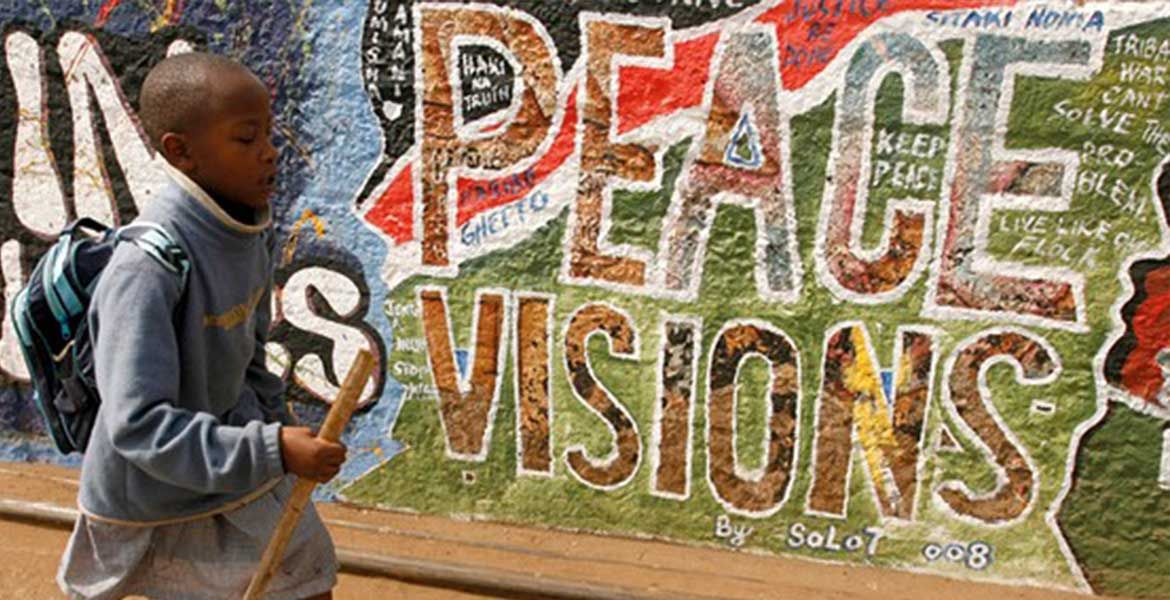 Ein Junge läuft mit einem Stock an einer Grafitti-Wand vorbei. Auf der Wand steht in großen Buchstaben "Peace Visions".