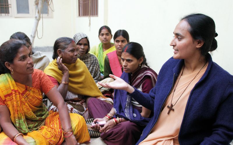 Eine junge Ordensschwester aus Indien spricht mit einer Frau in buntem Gewand, die ihr gegenüber auf dem Boden sitzt. Neben ihnen weitere Frauen unterschiedlichen Alters.