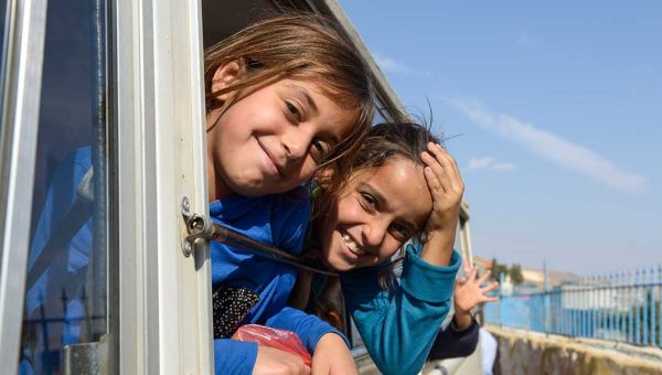 Zwei Mädchen schauen aus einem Busfenster und lachen in die Kamera.