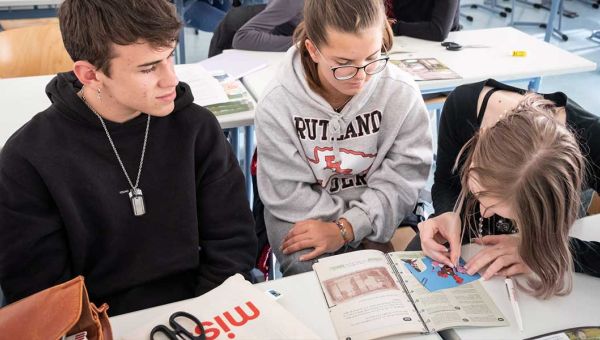 Ein Schüler und zwei Schülerinnen sitzen an einer Schulbank und sind in ein Arbeitsheft vertieft.
