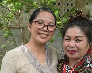 Zwei Frauen aus Mindanao/Philippinen lächeln vor einer Hauswand mit Bäumen in die Kamera: links Projektleiterin Jocelyn Aquiatan und rechts eine Begünstigte.