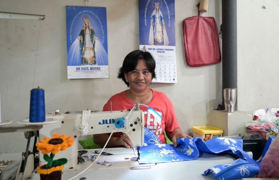 Eine Frau aus den Philippinen sitzt im Armenviertel Payatas in einem Raum an einem Tisch. Vor sich eine Nähmaschine und näht. Hinter ihr an der Wand zwei Kalender mit der heiligen Mutter Maria.