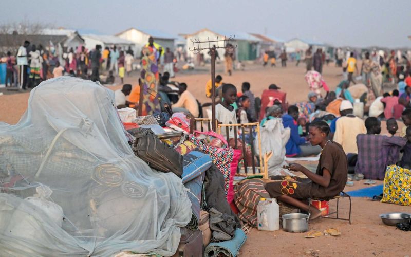 Viele Menschen sitzen unter freiem Himmel in der Abenddämmerung auf einer Straße im Camp, neben sich ihr Hab und Gut.