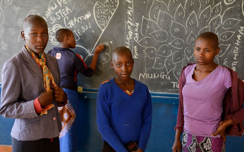 Vier junge Frauen stehen vor einer Schultafel. Drei von ihnen schauen in die Kamera, eine steht hinten an der Tafel und schreibt etwas mit Kreide darauf.