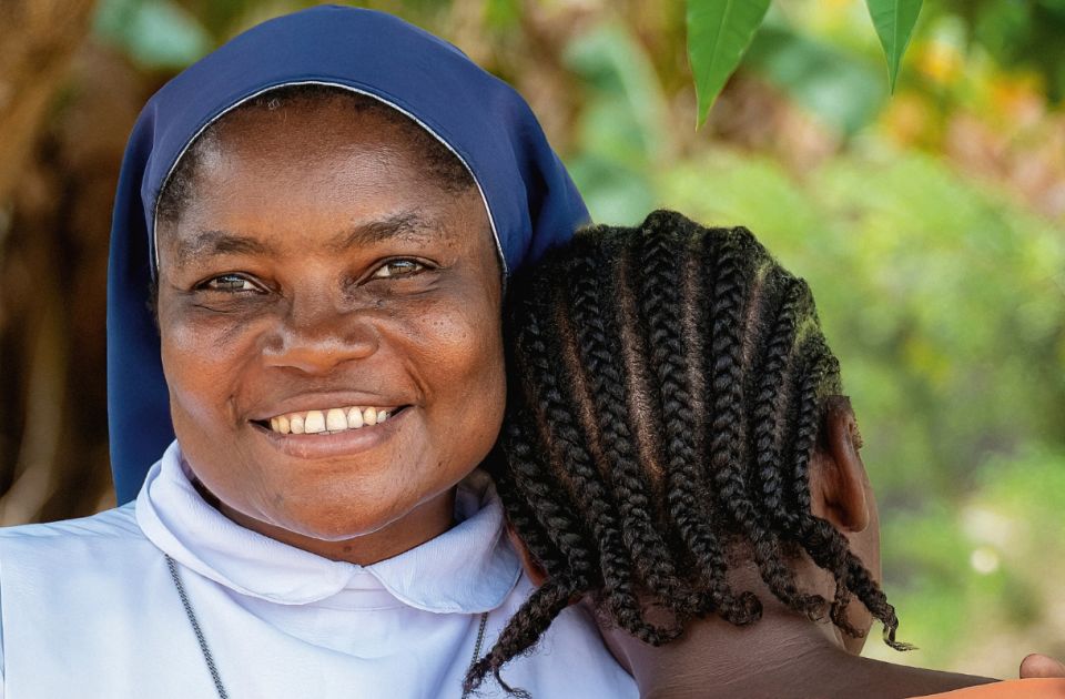 Afrikatag 2027: Eine Ordensschwester mit blauem Schleier und weißem Gewand aus Sierra Leone legt schützend ihre Hände auf die Schultern einer Frau in orangefarbener Kleidung, die dem Betrachter den Rücken zuwendet und ihre Wange an die linke Schulter der Ordensfrau lehnt.