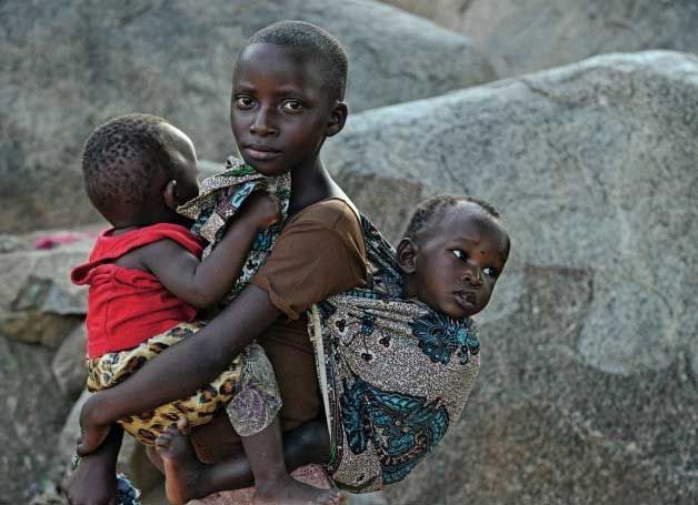 Eine Jugendliche trägt in einem Tragetuch auf dem Rücken ihr kleines Geschwister. Vorne hat sie ein weiteres Geschwister auf den Armt genommen.