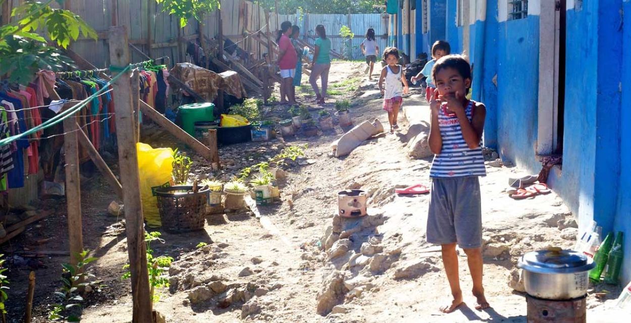 In einem Armenviertel von Cebu auf den Philippinen steht ein Kind im Vordergrund. Vor ihm auf dem Boden ein Topf auf einer offenen Feuerstelle, Kleider hängen auf Kleiderstangen auf der Seite des Weges.