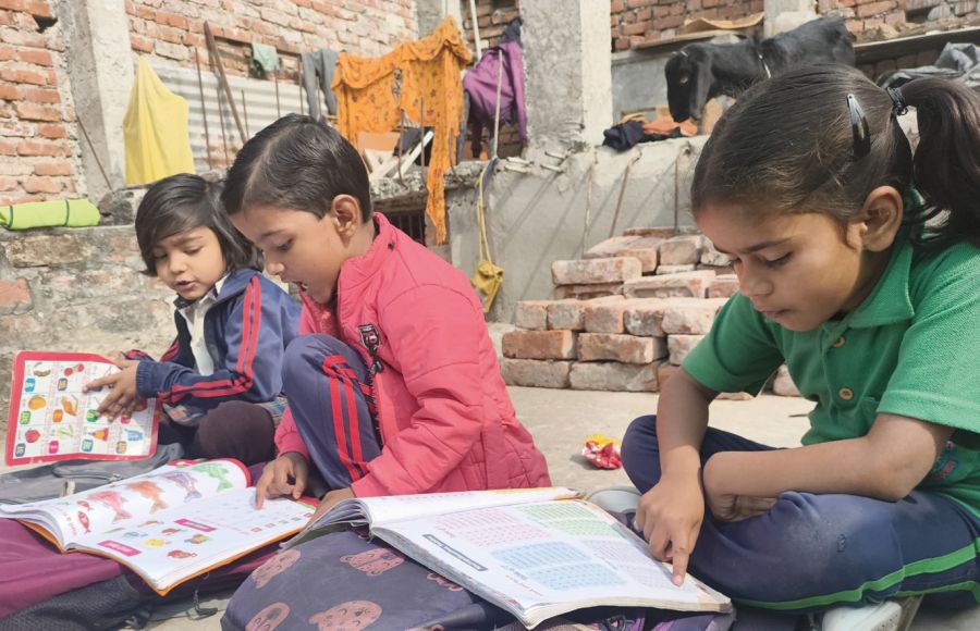 Drei indische Kinder im Grundschulalter sitzen auf dem Boden und blicken in ihre Schulhefte.