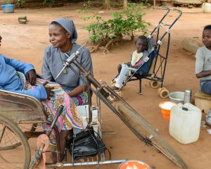 Eine Ordensfrau sitzt in einem Hof bei einer Frau im Rollstuhl und hört ihr zu. Im Hintergrund ist ein Kind in einem Buggy zu sehen und ein größeres Kind sitzt an einer Kochstelle.
