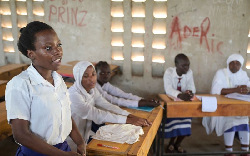 Schülerinnen an der Blue State Secondary des JRS in Kenia sitzen in einem Klassenzimmer. Ein Mädchen im Vordergrund steht und spricht in Richtung Tafel nach vorne.