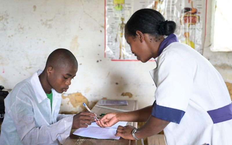 Ein jugendlicher Schüler in weißem Hemd beugt sich über ein Schulheft. Die Lehrerin wendet sich ihm zu, um ihm etwas zu zeigen.
