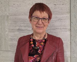 Porträtbild von Marita Krauss, die vor einer Wand aus Stein steht. rechts und links wurde der Hintergrund mit KI erweitert, um das erforderliche Bildformat zu erhalten.