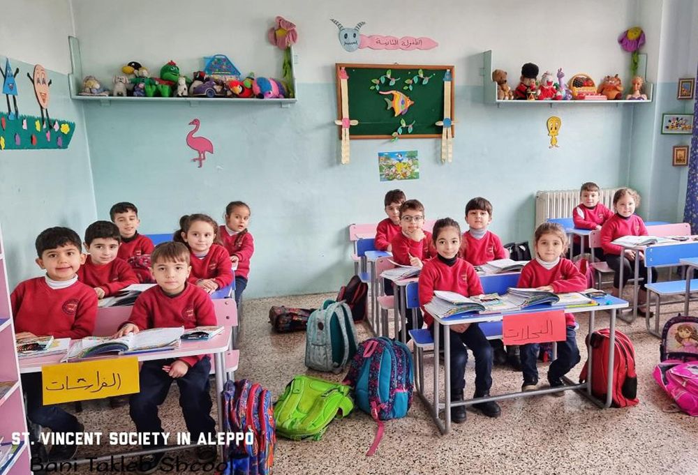 13 Mädchen und Jungen einer Grundschulklasse in Aleppo/Syrien sitzen in ihrem Klassenzimmer an Schulbänken und lacheln in die Karmera. Alle Schülerinnen und Schüler tragen einen roten Pullover und haben Schulbücher und Schulhefte vor sich liegen.