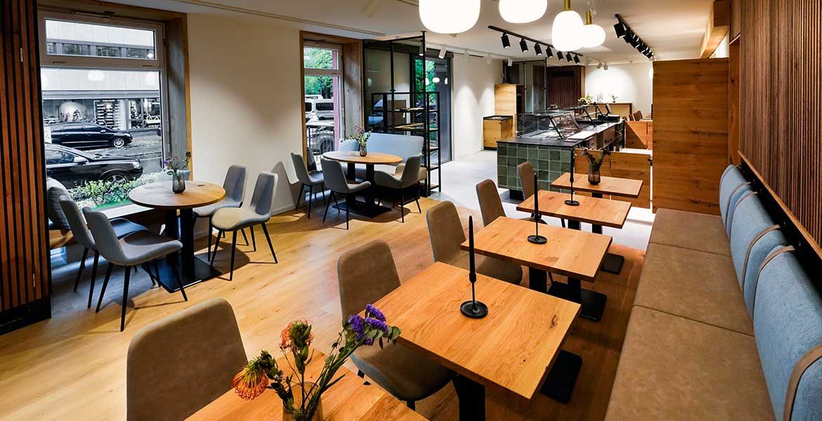 Innenansicht der Cafébar Conviva im Erdgeschoss bei missio München. Blick durch den langegezogenen Raum über die Sitzgruppen in Richtung der Fenster zur Straße.