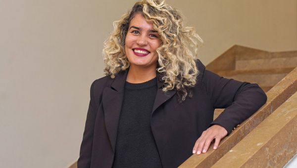Porträt von Suzannah Mirghani, die auf einer Treppe sitzt und in die Kamera lächelt. Das Schwarz ihrer Kleidung kontrastiert mit ihren blond gefärbten Locken und dem bordeauxroten Lippenstift.