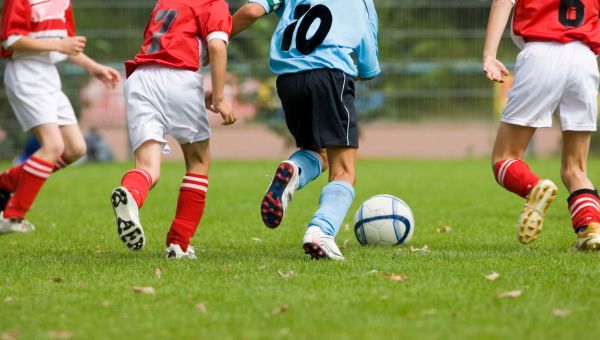 Auf einem Rasenplatz liegt ein Fußball. Vier Jungen bewegen sich auf den Ball zu. Zu sehen sind nur Beine und Rumpf. Drei Jungs tragen ein rotes Trikot mit weißer Hose, ein weiterer Junge ein blaues Trikot mit schwarzer Hose.