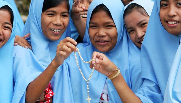 Junge Frauen mit hellblauen Tüchern um Kopf und Schultern halten einen Rosenkranz in die Kamera und lächeln.