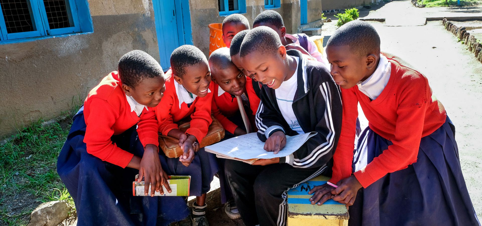 Kinder in Schuluniform sind um einen Jungen gruppiert, der aus einem Heft vorliest. Die Kinder aus Kenia lauschen mit neugierigem, freudigen Gesichtsausdruck 