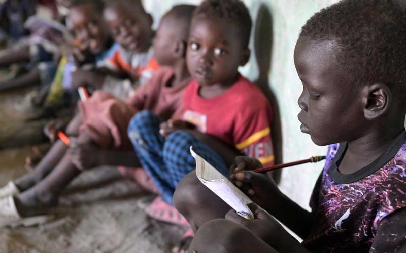 Im Vordergrund rechts im Bild sitzt ein Junge aus dem Südsudan auf dem Boden und schreibt mit einem Bleistift konzentriert in ein Heft. Im Hintergrund neben ihm sind weitere Kinder zu sehen, die zu ihm blicken.