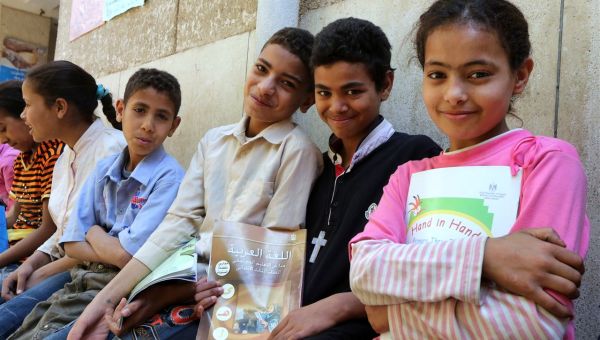 Sechs arabische Kinder sitzen nebeneinander auf einer Bank. Ein Junge trägt ein Kreuz um den Hals, ein Mädchen hält eine deutschsprachige Broschüre in der Hand.
