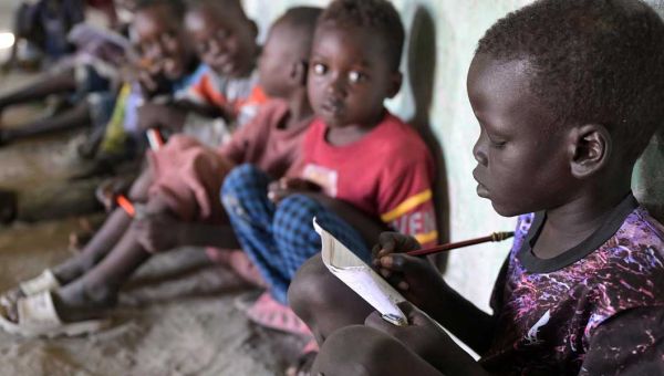 Im Vordergrund rechts im Bild sitzt ein Junge aus dem Südsudan auf dem Boden und schreibt mit einem Bleistift konzentriert in ein Heft. Im Hintergrund neben ihm sind weitere Kinder zu sehen, die zu ihm blicken.