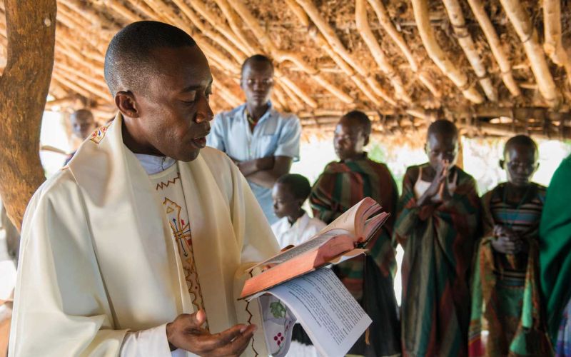 Ein Priester steht in Uganda unter einer Überdachung aus Holz und Lehm und liest aus einer aufgeschlagenen Bibel. Im Hinjtergrund sieht man betende Gläubige.
