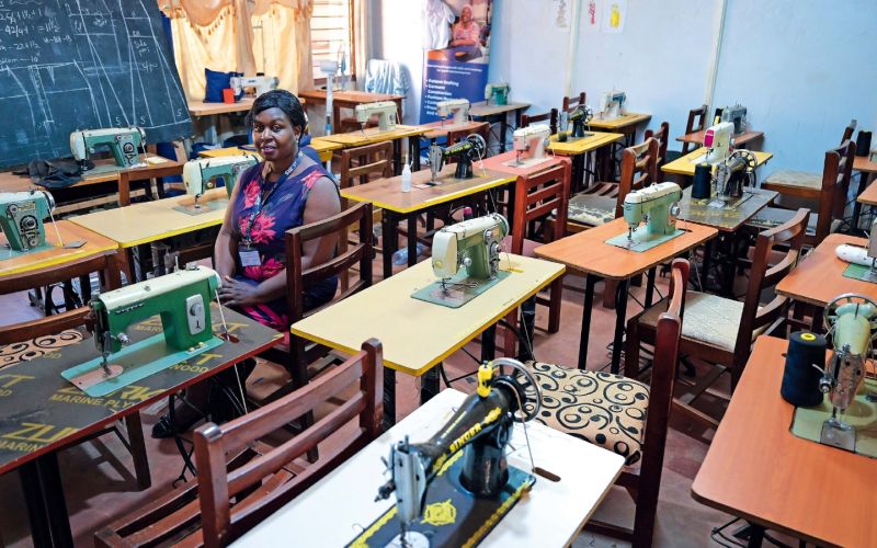 Eine Frau sitzt allein im Schulungsraum, vorne eine Tafel mit Schnittmustern, auf den Tischen stehen Nähmaschinen. Davor je ein leerer Stuhl.