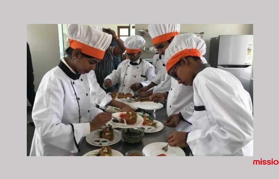 Vier ehemalige Straßenkinder stehen in weißer Kochuniform und Kochmützen in einer Küche und dekorieren Teller mit Essen.