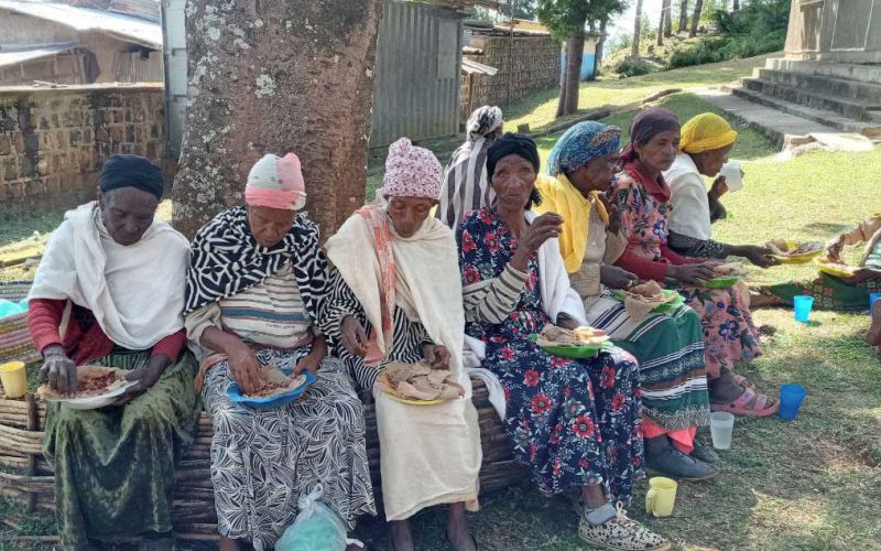 Eine Gruppe älterer Frauen isst auf einer Bank vor einem Baum mit der Hand äthiopische Teigfladen.