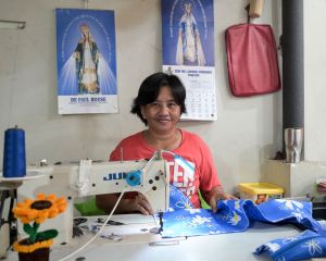 Eine Frau aus den Philippinen sitzt im Armenviertel Payatas in einem Raum an einem Tisch. Vor sich eine Nähmaschine und näht. Hinter ihr an der Wand zwei Kalender mit der heiligen Mutter Maria.
