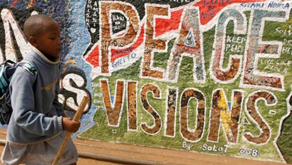 Ein Junge läuft mit einem Stock an einer Grafitti-Wand vorbei. Auf der Wand steht in großen Buchstaben "Peace Visions".