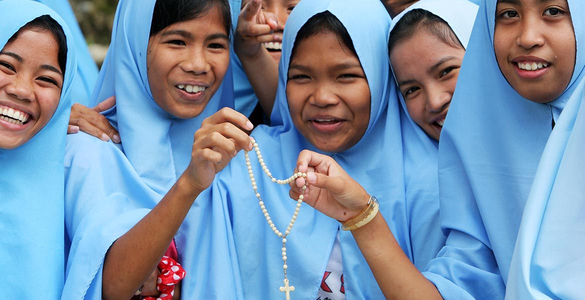Junge Frauen mit hellblauen Tüchern um Kopf und Schultern halten einen Rosenkranz in die Kamera und lächeln.