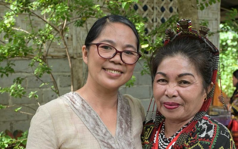 Zwei Frauen aus Mindanao/Philippinen lächeln vor einer Hauswand mit Bäumen in die Kamera: links Projektleiterin Jocelyn Aquiatan und rechts eine Begünstigte.