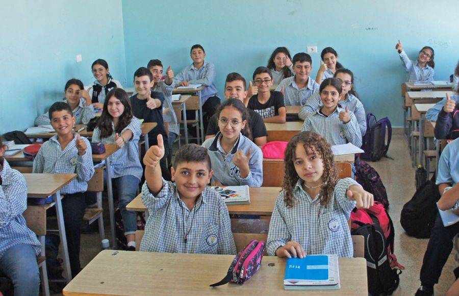 Schulkinder aus dem Libanon sitzen in hellblauer Schuluniform in einem Klassenzimmer an Schultischen. Sie lachen in die Kamera, einige von ihnen strecken ihren Daumen nach oben.
