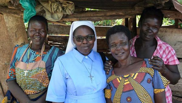 Vier Frauen aus Uganda stehen unter vor einer Bretterwand mit Dach und lächeln in die Kamera. Die zweite von links ist eine Ordensschwester.