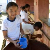 Ein madagassisches Mädchen hält einen blauen Kunststoffbecher in der Hand und lächelt. Vor ihr eine Tonne mit Trinkwasser. Im Hintergrund sind mehrere Kinder zu sehen, die entlang der Wasserleitung stehen und trinken oder sich die Hände waschen.