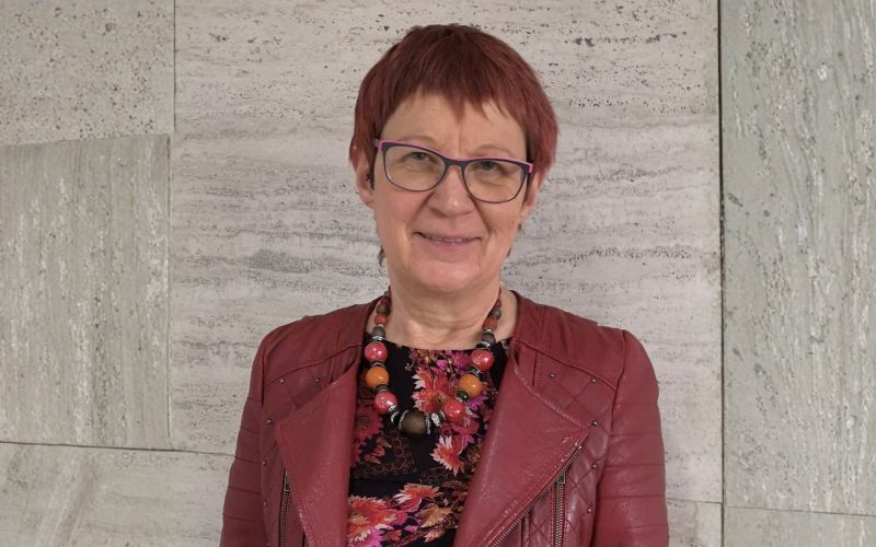 Porträtbild von Marita Krauss, die vor einer Wand aus Stein steht. rechts und links wurde der Hintergrund mit KI erweitert, um das erforderliche Bildformat zu erhalten.