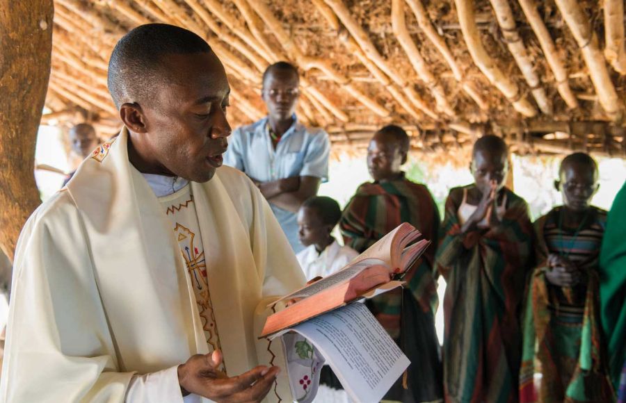 Ein Priester steht in Uganda unter einer Überdachung aus Holz und Lehm und liest aus einer aufgeschlagenen Bibel. Im Hinjtergrund sieht man betende Gläubige.