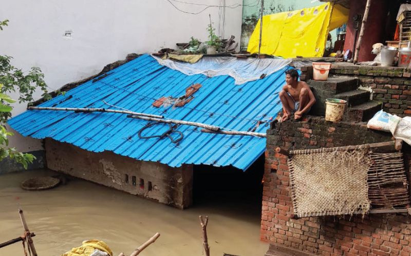 Flut in Indien: Ein Mann sitzt erhöht auf einer Mauer neben einer Wellblechhütte, die zu großen Teilen bereits überschwemmt ist.