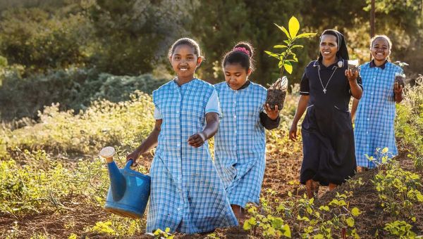 Eine Ordensfrau und drei Mädchen laufen in Madagaskar über ein Feld auf die Kamera zu. Das erste Mädchen trägt in der rechten Hand eine Gießkanne, das hinter ihr laufende Mädchen, die Ordensschwester dahinter und das letzte Mädchen tragen jeweils einen Pflanzensetzling in ihren linken Händen. Alle vier lächeln.