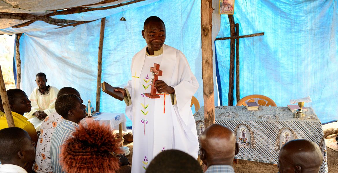 Ein afrikanischer Priester steht vor der Gemeinde  in einem provisorischen Kirchenraum.