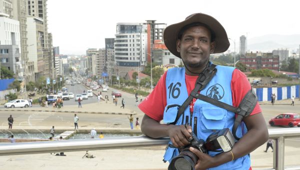 Porträt von Adana Fanti mit seiner Kamera, im Hintergrund eine vielbefahrene Kreuzung und ein Platz mit Brunnen in Addis Abeba/Äthiopien.