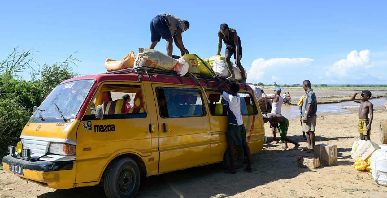 Das Dach eines gelben Kleinbusses wird in Madagaskar auf sandigem Boden von mehreren Männern mit Säcken beladen.