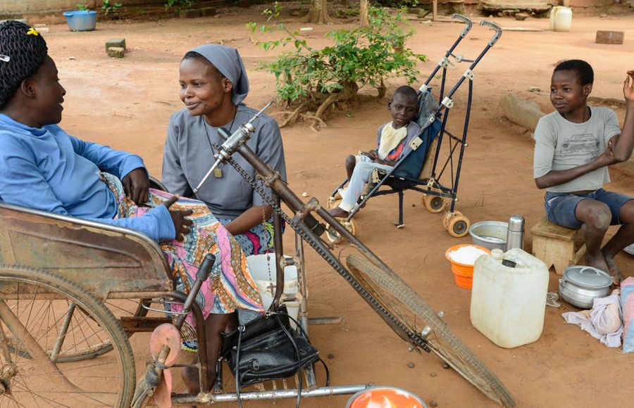 Eine Ordensfrau sitzt in einem Hof bei einer Frau im Rollstuhl und hört ihr zu. Im Hintergrund ist ein Kind in einem Buggy zu sehen und ein größeres Kind sitzt an einer Kochstelle.