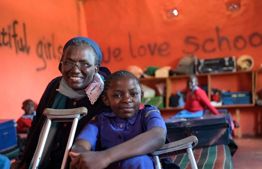 In einem Klassenzimmer mit rot-orangenen Wänden ist im Vordergrund eine Ordensschwester aus Kenia zu sehen, die neben einem Mädchen mit Behinderung sitzt. Vor der Ordensschwester lehnt die Krücke des Mädchens.